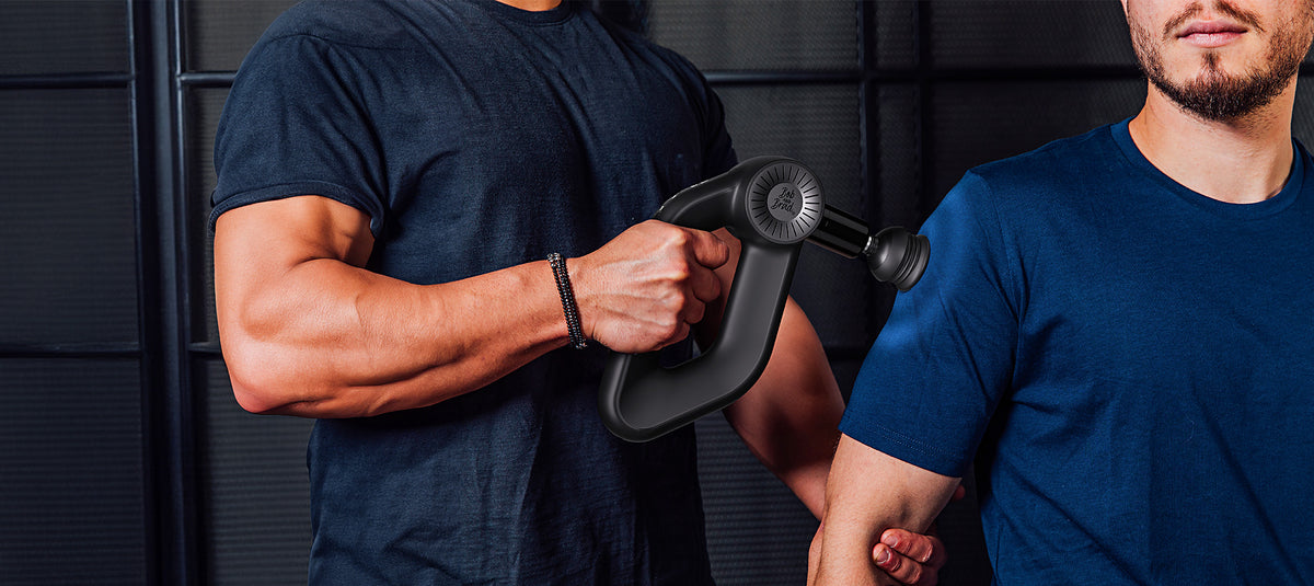A man in a navy blue t-shirt holds a black handheld massage gun against the upper arm of another man wearing a blue t-shirt, set against a dark paneled background.