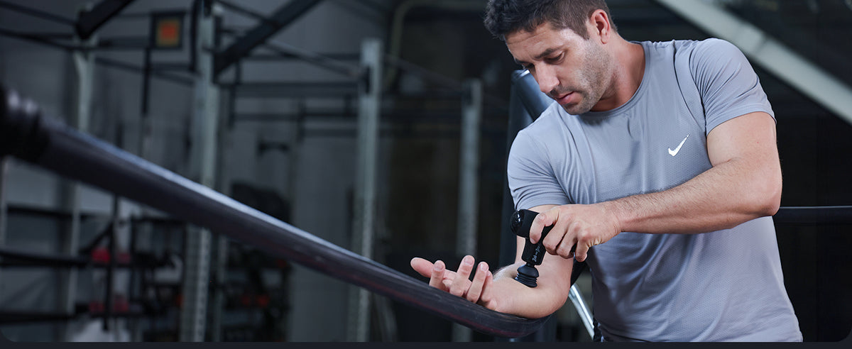 A man in a gray Nike athletic T-shirt is shown in a gym setting, holding a Q2 massage gun to massage his forearm. The image is in a gym-like setting.