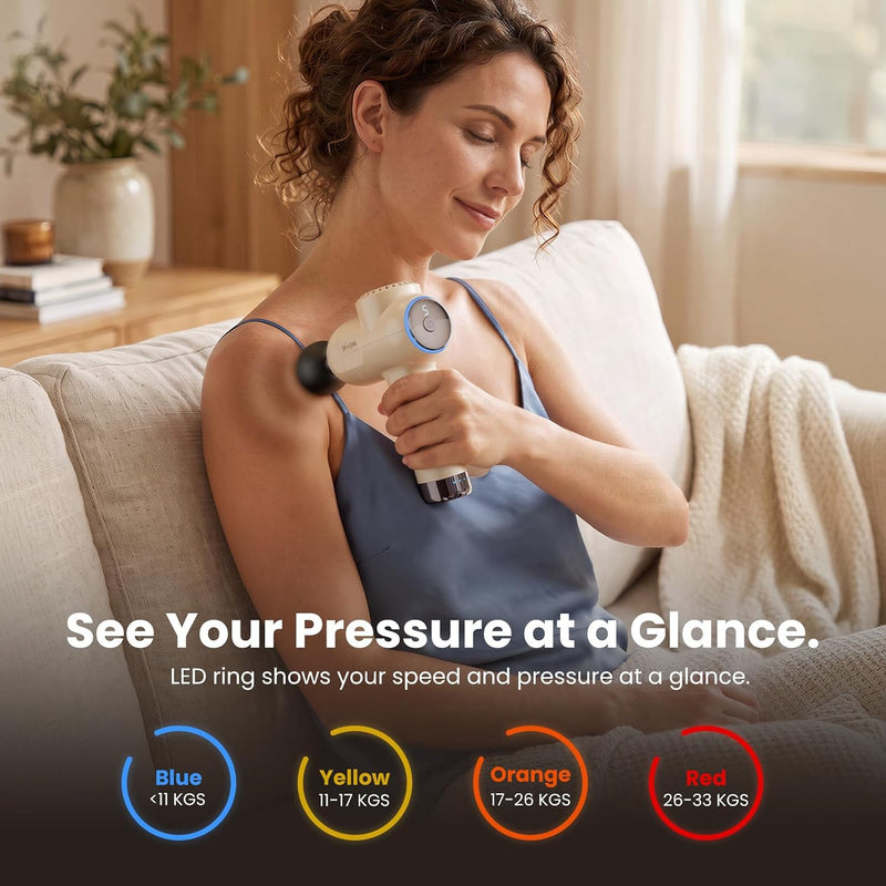 Woman using the Bob and Brad C2 Ultra Massage Gun on her shoulder while seated on a couch. Color-coded LED pressure indicators show blue (under 11 kg), yellow (11–17 kg), orange (17–26 kg), and red (26–33 kg).