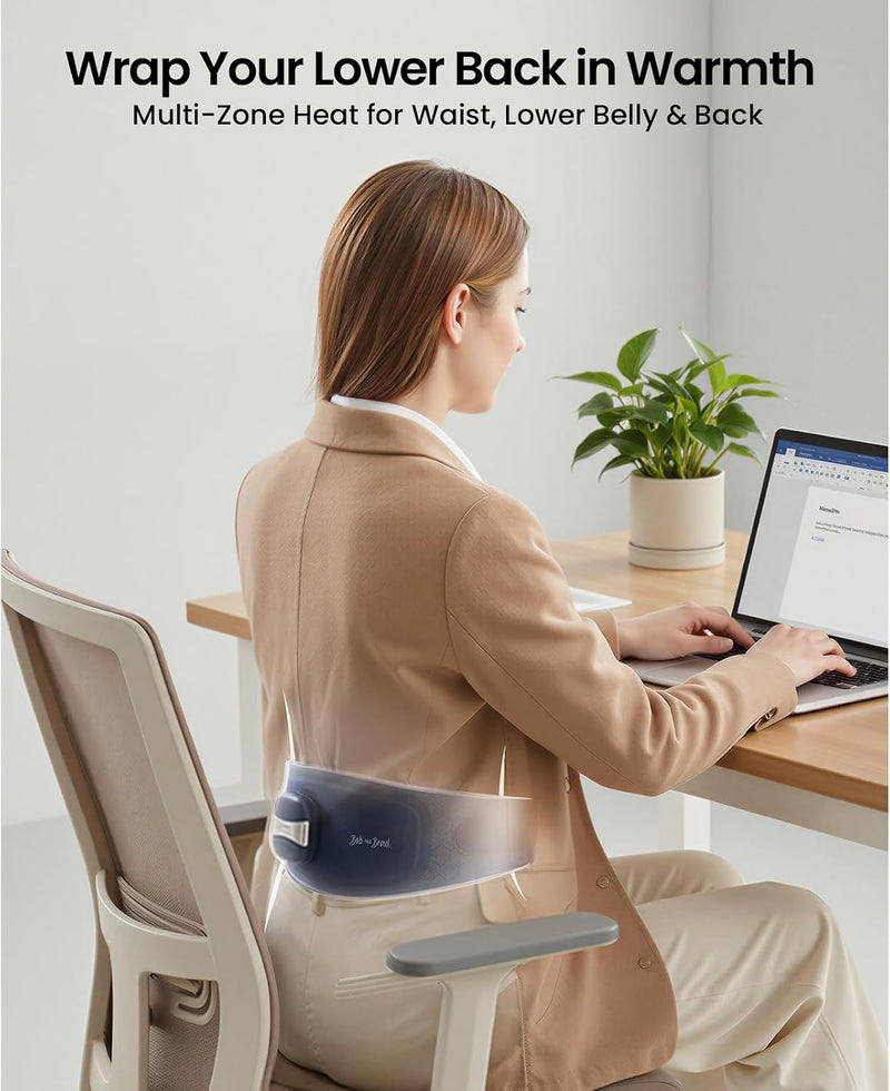 A woman sits at a desk using a lumbar back heated wrap around her lower back. The scene includes text at the top reading 'Wrap Your Lower Back in Warmth' and 'Multi-Zone Heat for Waist, Lower Belly & Back,' with a chair, laptop, and potted plant in a bright indoor setting.