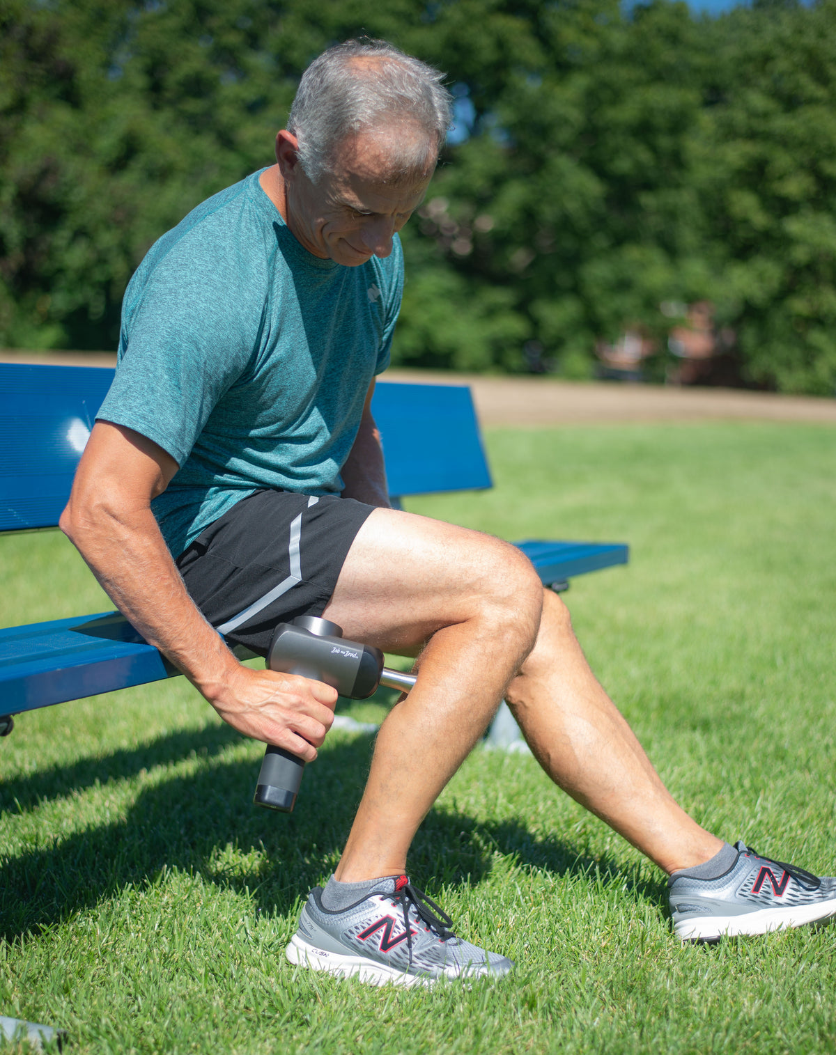 Brad is sitting on a blue park bench outdoors, using a handheld massage gun on his leg. He is wearing a teal t-shirt, black athletic shorts, and gray New Balance sneakers on a green grassy field with trees in the background.