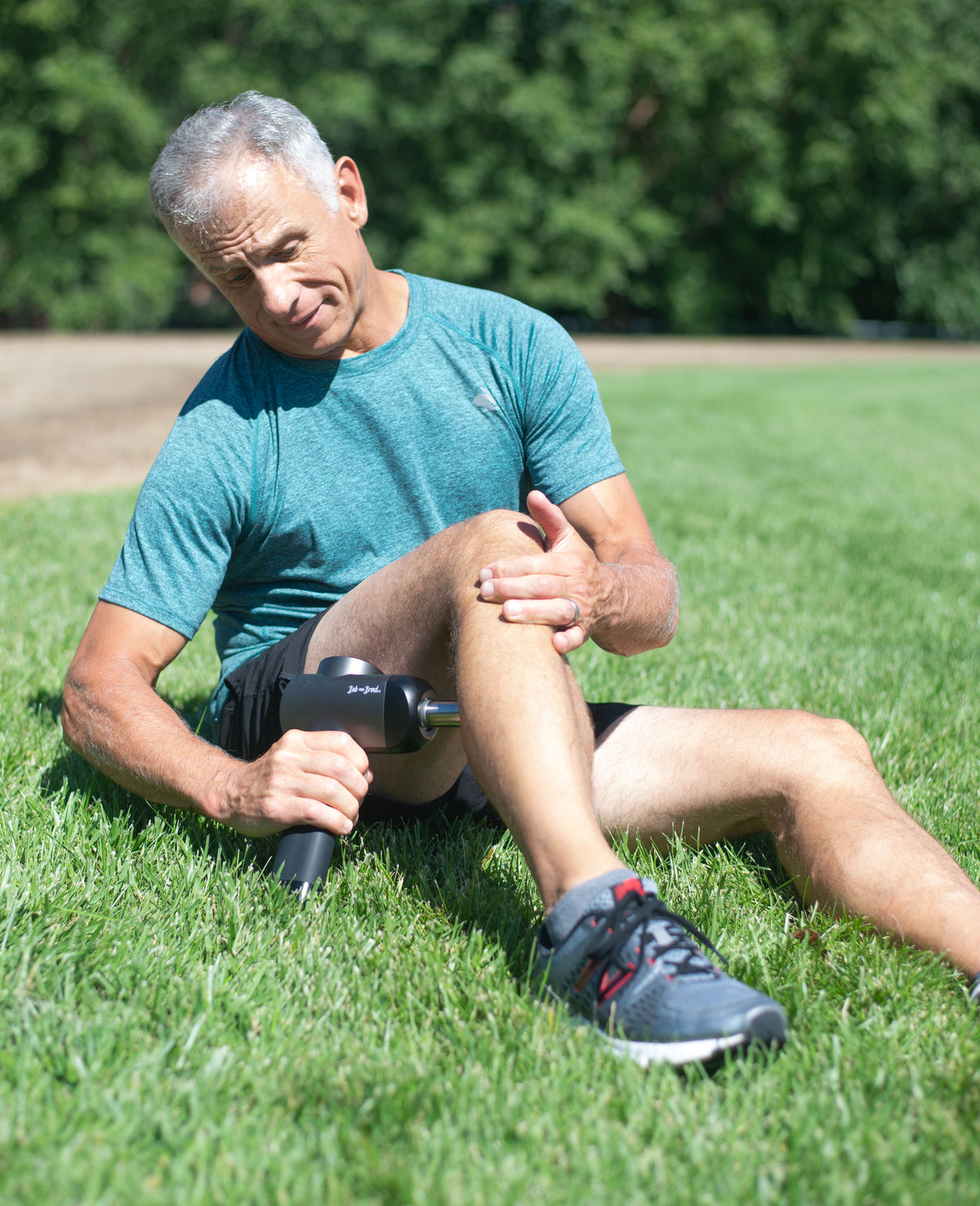 Brad, sitting on green grass in a park uses a handheld percussion massage gun on his leg. He is wearing a teal athletic t-shirt, black shorts, and gray running shoes.