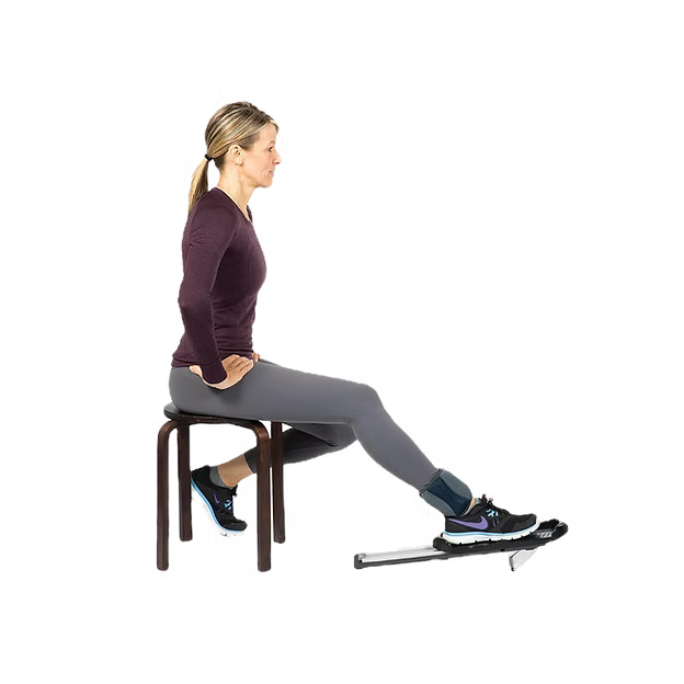 A woman in profile sits on a dark wooden stool, wearing gray leggings, a dark plum long-sleeve top, and athletic shoes, with a black and silver Bob and Brad Knee Glide device under her right foot and an ankle weight around her ankle.