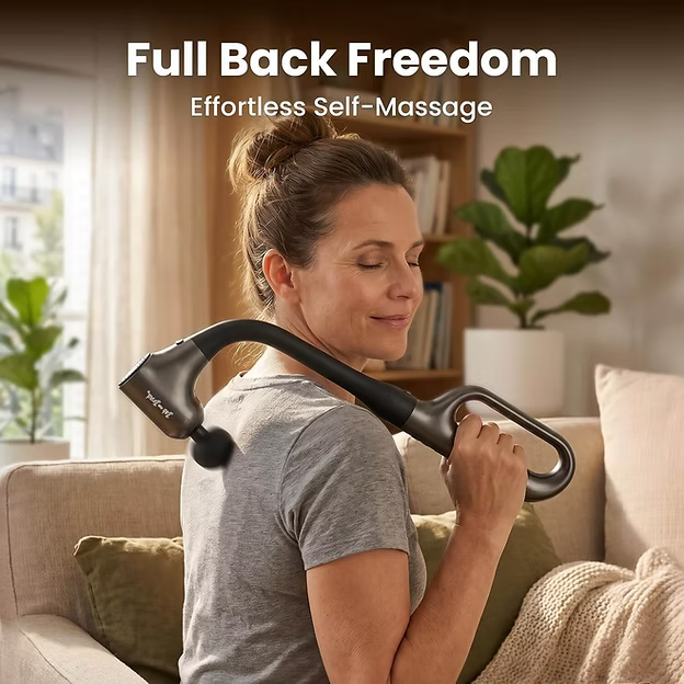 A woman sitting on a beige couch in a living room uses a dark gray, long-handled handheld massage gun on her upper shoulder. Text at the top reads 'Full Back Freedom' and 'Effortless Self-Massage'.