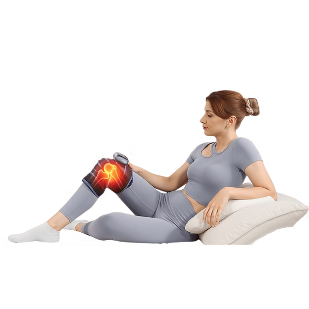 A woman in a gray workout set reclines on a white pillow while using a heated knee massage device on her right knee. The device displays a glowing red and orange heat graphic to indicate massage or heat function, set against a plain white background.
