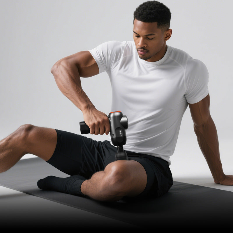 A man in a white t-shirt and black shorts sits on a black exercise mat using a Bob and Brad handheld percussion massage gun on his upper leg, against a light gray studio background.