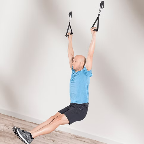 A man in a blue T-shirt and black shorts hangs using black handles attached to a wall with wall anchors in a minimalist room with a light wood floor.