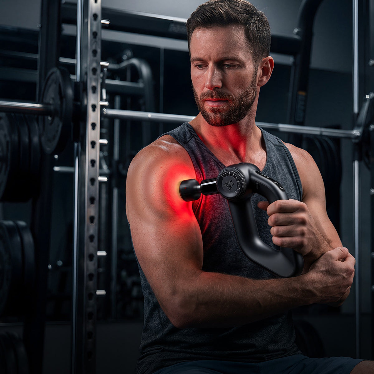 Man using the Bob and Brad D6 massage gun on his bicep in a gym setting. A red glow coming from the massage head. 