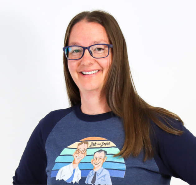Adrienne, Doctor of Physical Therapy, with long brown hair and glasses, wearing a navy blue Bob & Brad T-shirt with a colorful graphic print on the front, set against a plain white background.