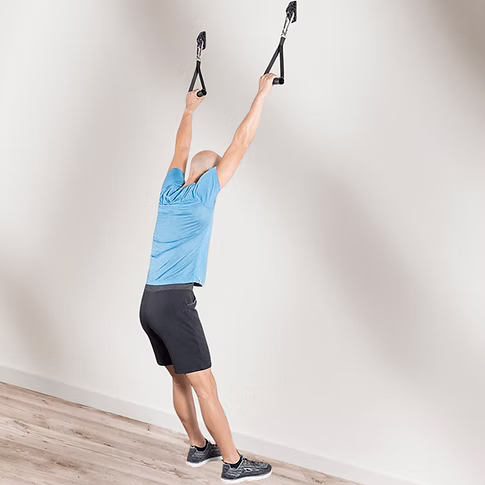 A man in a blue T-shirt and black shorts performs a pull-up exercise using black handles attached to a wall with wall anchors in a minimalist room with a light wood floor.