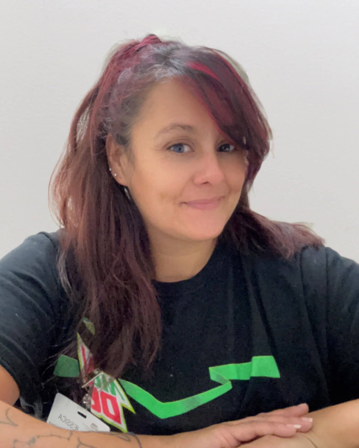 Jessie with dark reddish-brown hair wearing a black T-shirt with a green graphic logo, looking directly at the camera against a plain light gray wall.