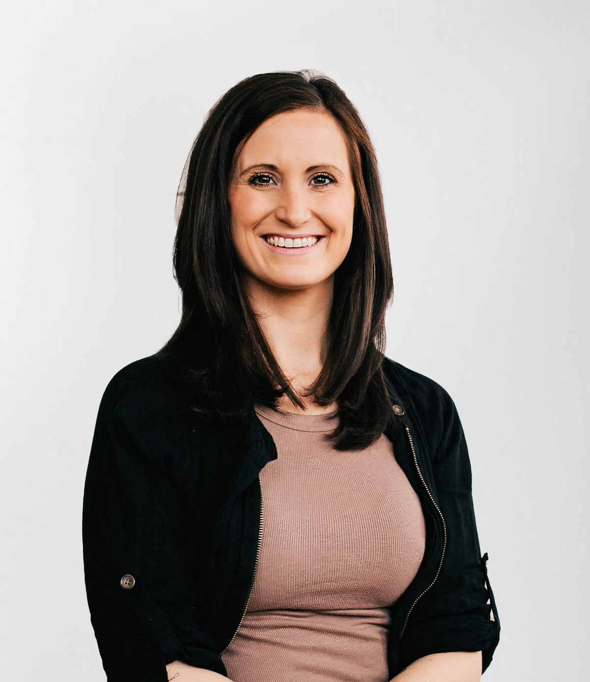 Liz with dark brown hair wearing a black zip-up jacket over a dusty rose ribbed top, photographed against a plain light gray background.