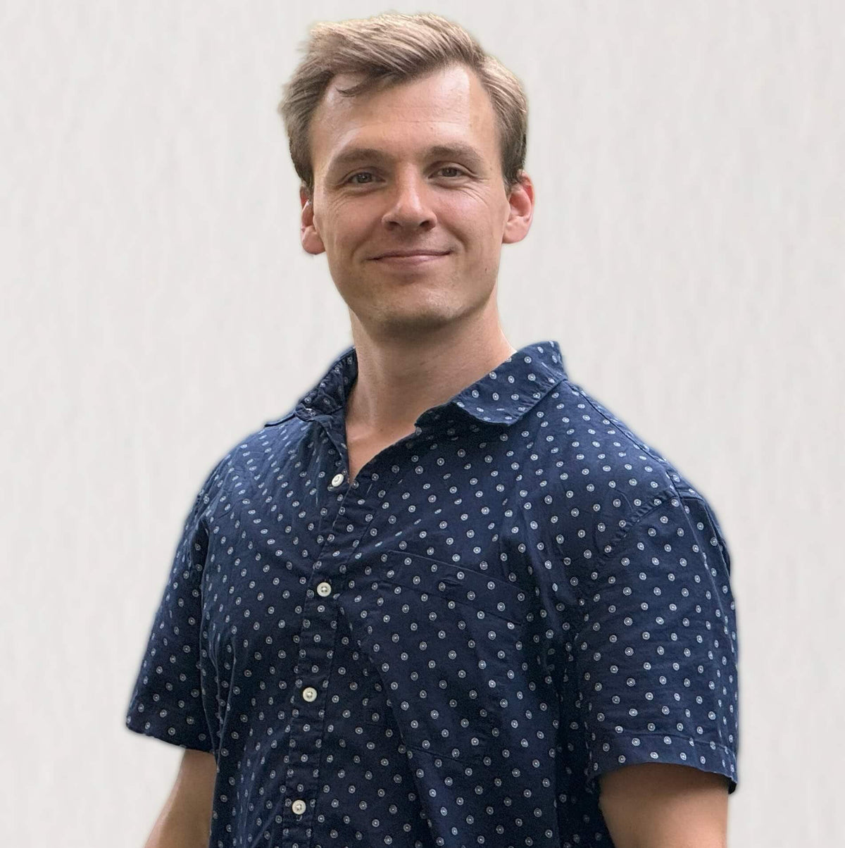 Matt is wearing a navy blue short-sleeve button-up shirt with a white polka dot pattern, photographed against a light gray textured background.