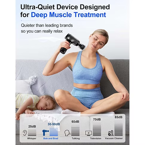 A woman in a blue sports bra and matching shorts sits cross-legged on a gray sofa while using a black handheld massage gun on her shoulder. A child is sleeping on the couch beside her.