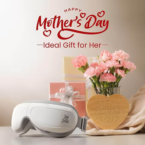 A white and gray eye massager is displayed on a tabletop alongside pink carnations in a vase, a wooden heart-shaped plaque reading 'Happy Mother's Day,' gift boxes, and a beige cloth. Red text at the top reads 'Happy Mother's Day' and 'Ideal Gift for Her.