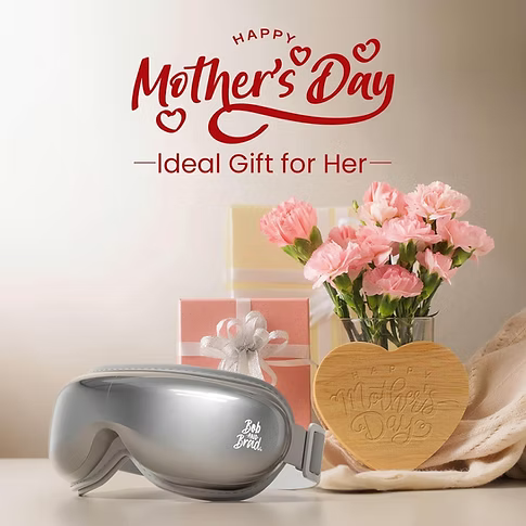 Silver eye massager gift set on a light beige surface with pink carnations in a glass vase, a wooden heart-shaped Mother's Day plaque, wrapped gift boxes, and a red 'Happy Mother's Day' sign above.