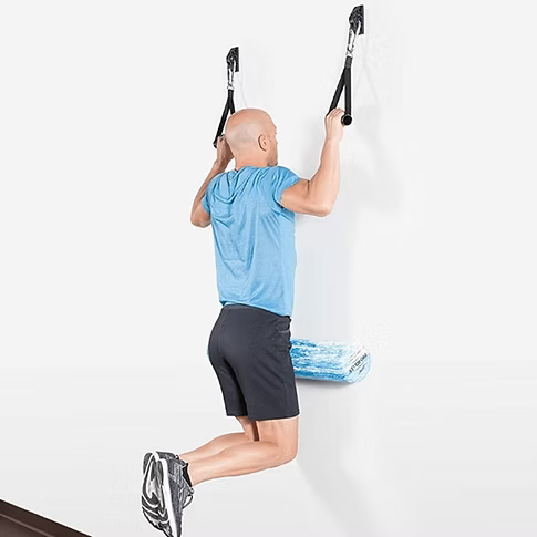 A man performing a wall hang exercise using black handles attached to a white wall, with a blue foam roller between him and the wall. He is wearing a light blue T-shirt, dark gray shorts, and athletic shoes.