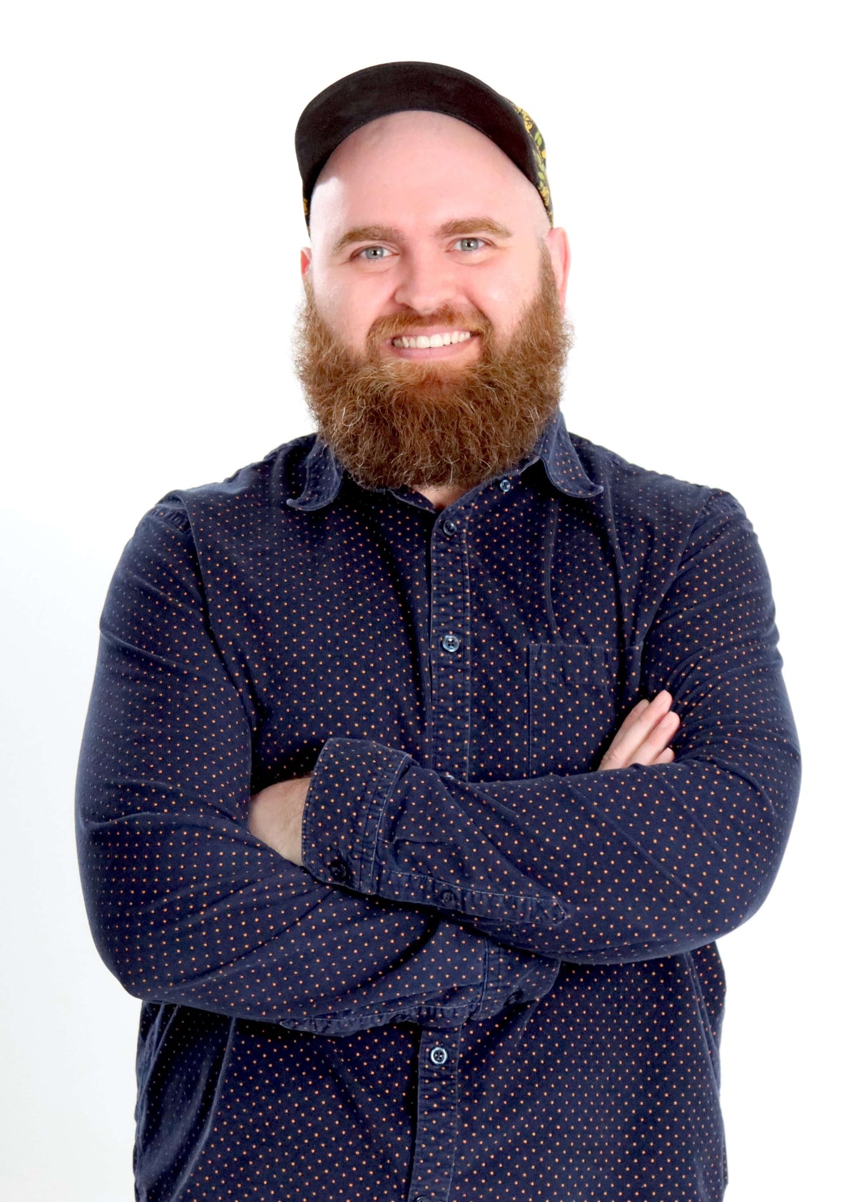 Tanner, with a full red beard and a black skullcap stands with arms crossed, wearing a dark navy long-sleeve button-up shirt with a small white polka dot pattern, against a plain white background.