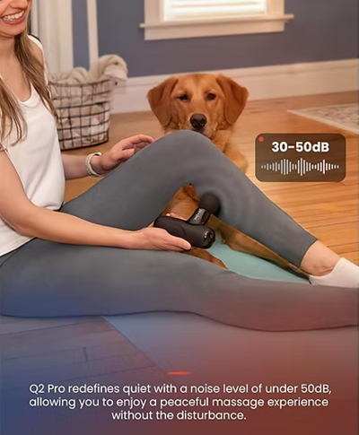 A woman in gray leggings sits on a wooden floor indoors while using a small black massage device on her leg, with a golden retriever beside her. A text badge says '30- 50 dB '. Text includes: Q2 Pro redefines quiet with a noise level of under 50dB, allowing you to enjoy a peaceful massage experience without the disturbance. 