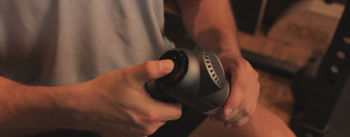 Load video: A man in a gym setting presses a button on the back of a massage gun to start it and then proceeds to use the massage gun to massage his biceps and quads. 
