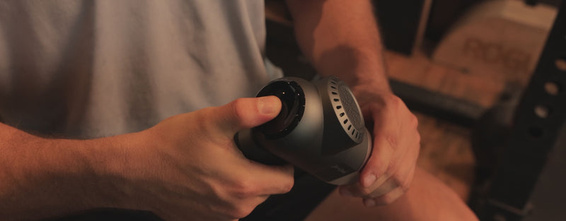 A man in a gym setting presses a button on the back of a massage gun to start it and then proceeds to use the massage gun to massage his biceps and quads. 